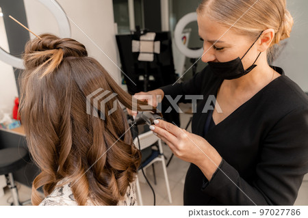 A hairdresser in a black mask prepares a woman, makes a hairstyle with curls using a curling iron. Long light brown natural hair. Self care concept. 97027786