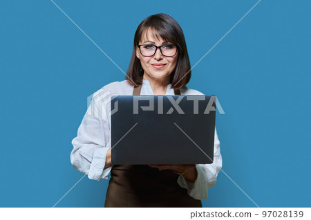 Portrait of middle aged woman in apron with laptop on blue background. 97028139