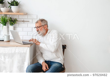 Handsome old man dressed in hoodie and eyeglasses is using laptop and smiling while listen music sitting on couch at home copy space and empty place for text 97028482