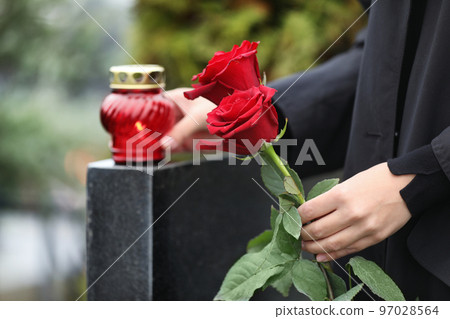 Woman holding red roses near black granite tombstone with candle outdoors, closeup. Funeral ceremony Woman holding red roses near black granite tombstone with candle outdoors, closeup. Funeral ceremony 97028564