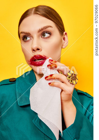 Young woman in green coat with smudged red lipstick wiping mouth after eating nuggets over yellow background. Food pop art photography Young woman in green coat with smudged red lipstick wiping mouth after eating nuggets over yellow background. Food pop art photography 97029066