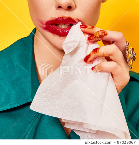 Cropped image of woman in green coat with smudged red lipstick cleaning mouth after eating nuggets over yellow background. Food pop art photography. 97029067