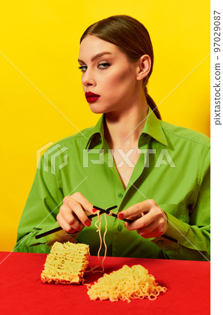 Woman with emotionless face knitting instant noodles on blue table on vivid red tablecloth over yellow background. Food pop art photography. Woman with emotionless face knitting instant noodles on blue table on vivid red tablecloth over yellow background. Food pop art photography. 97029087