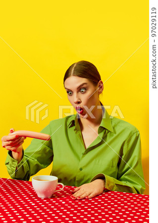 Emotional young woman vintage clothes eating sausage with coffee, cappuccino on red tablecloth over yellow background. Food pop art photography. 97029119