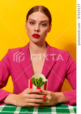Emotionless, serious young woman in bright pink jacket holding green beans over yellow background. Healthy diet. Food pop art photography. 97029137