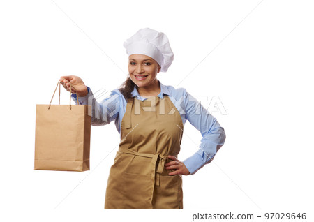 Adorable woman waitress or chef confectioner, baker in blue casual shirt and beige apron, smiling and holding out a disposable eco paper bag with fresh delivered food, isolated over white background 97029646