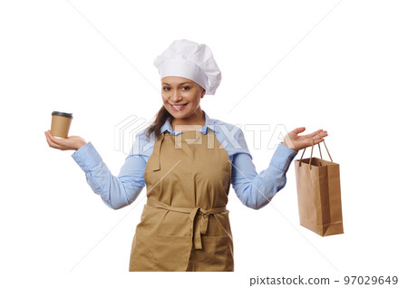 Cute woman waitress, bartender, confectioner wearing chef's hat and apron, smiling a cheerful toothy smile, looking at camera, holding a cardboard cup of takeaway hot tea or coffee, isolated on white 97029649