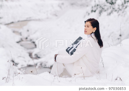 A stylish woman with a white suit with a New Year's gift in her hands in a winter forest. A girl in nature in a snowy forest with a gift box A stylish woman with a white suit with a New Year's gift in her hands in a winter forest. A girl in nature in a snowy forest with a gift box 97030853