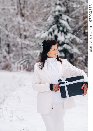 A stylish woman with a white suit with a New Year's gift in her hands in a winter forest. A girl in nature in a snowy forest with a gift box 97030858
