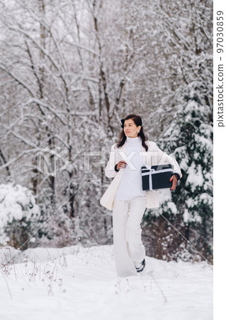 A stylish woman with a white suit with a New Year's gift in her hands in a winter forest. A girl in nature in a snowy forest with a gift box 97030859