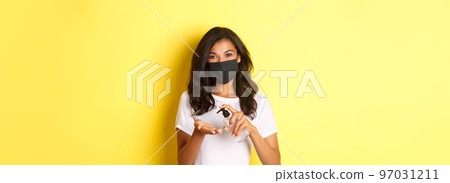 Concept of covid-19, social distancing and lifestyle. Image of african-american girl in face mask using hand sanitizer, standing over yellow background Concept of covid-19, social distancing and lifestyle. Image of african-american girl in face mask using hand sanitizer, standing over yellow background 97031211