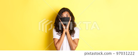 Concept of coronavirus, pandemic and lifestyle. Portrait of excited african-american woman in face mask, looking amazed at something cool, standing over yellow background Concept of coronavirus, pandemic and lifestyle. Portrait of excited african-american woman in face mask, looking amazed at something cool, standing over yellow background 97031222