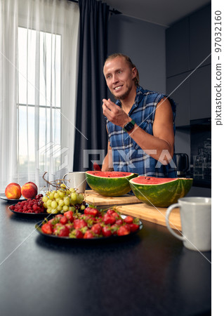 Handsome young sporty smiling man in the kitchen is preparing vegan healthy fruits salad and smoothie in a good mood Handsome young sporty smiling man in the kitchen is preparing vegan healthy fruits salad and smoothie in a good mood 97032160
