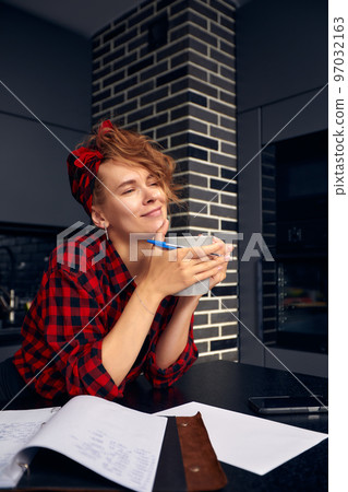 Joyful young female working from home, doing paperwork, having coffee break, sitting at dining table with mug and diary, making to do list. Cute girl sketching in copybook and drinking tea Joyful young female working from home, doing paperwork, having coffee break, sitting at dining table with mug and diary, making to do list. Cute girl sketching in copybook and drinking tea 97032163