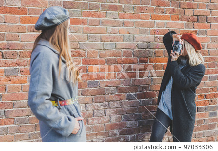 Girl takes picture of her female friend in front of brick wall in urban street - photographer and youth urban lifestyle concept Girl takes picture of her female friend in front of brick wall in urban street - photographer and youth urban lifestyle concept 97033306