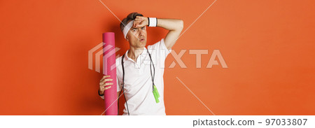 Concept of workout, gym and lifestyle. Image of handsome middle-aged man, tired after fitness exercises, holding skipping rope and yoga mat, wiping sweat off forehead, orange background 97033807