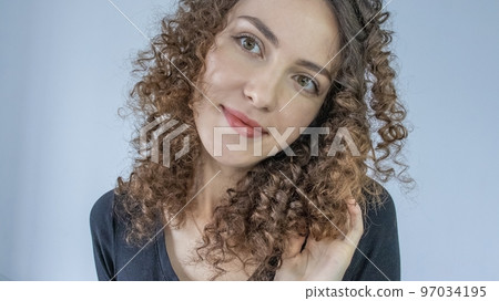 Headshot of a cheerful girl with curly hair in a black T-shirt with a smile, against a gray background. Perming a woman's hair 97034195