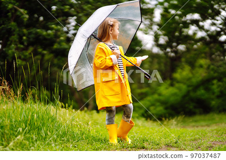 Little girl with transparent umbrella playing in the rain on sunny. Beautiful girl in a yellow cloak and rubber boots. 97037487