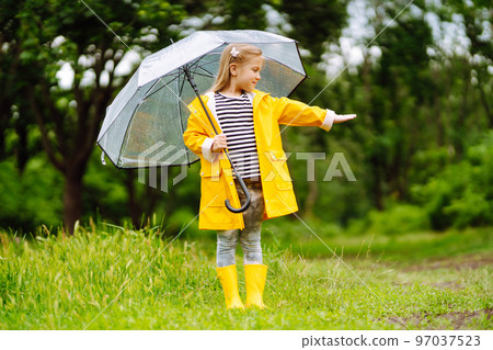 Little girl with transparent umbrella playing in the rain on sunny. Beautiful girl in a yellow cloak and rubber boots. 97037523