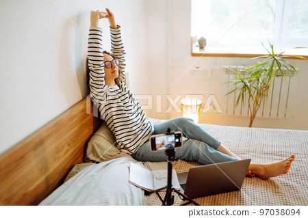 Young woman blogger talking with followers, live streaming, looking to phone screen sitting on bed. 97038094
