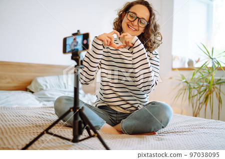 Young woman blogger talking with followers, live streaming, looking to phone screen sitting on bed. 97038095