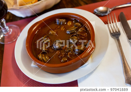 Stewed snails in red sauce served on a white ceramic plate close to the cutlery and bread in the restaurant 97041889