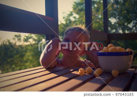 Little girl is sitting on porch in summer. Cheese balls in bowl. Cheese flavoured snacks on a table. Dreamy and romantic image. Summer and happy childhood concept Little girl is sitting on porch in summer. Cheese balls in bowl. Cheese flavoured snacks on a table. Dreamy and romantic image. Summer and happy childhood concept 97043185