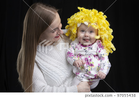 Mom holds in her arms a little happy daughter in a yellow wig. Funny kids. Mom holds in her arms a little happy daughter in a yellow wig. Funny kids. 97055438