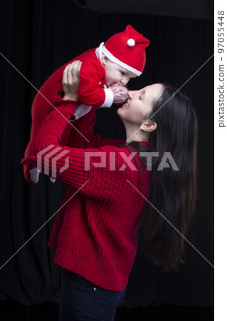 Mom and little child at Christmas. A woman holds her son in her arms, who is dressed in Santa Claus clothes. 97055448