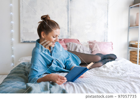 Leisure and people concept young woman reading book in bed in blue shirt Leisure and people concept young woman reading book in bed in blue shirt 97057735
