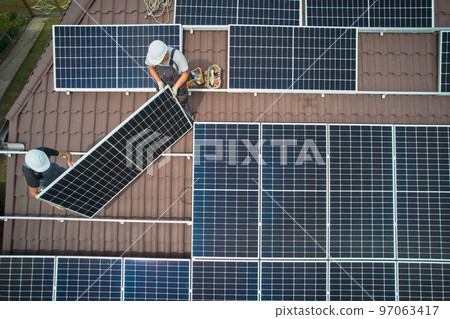 Men workers installing photovoltaic solar moduls on roof of house. Engineers in helmet building solar panel system outdoors. Concept of alternative and renewable energy. Aerial view. Men workers installing photovoltaic solar moduls on roof of house. Engineers in helmet building solar panel system outdoors. Concept of alternative and renewable energy. Aerial view. 97063417