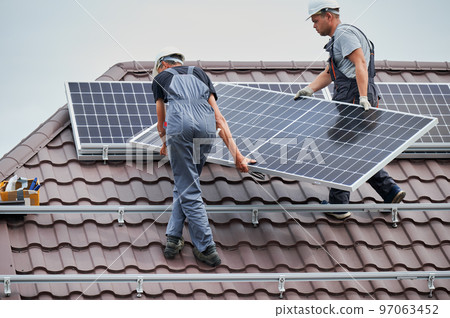 Men technicians carrying photovoltaic solar moduls on roof of house. Workmen in helmets mounting solar panel system outdoors. Concept of alternative and renewable energy. 97063452
