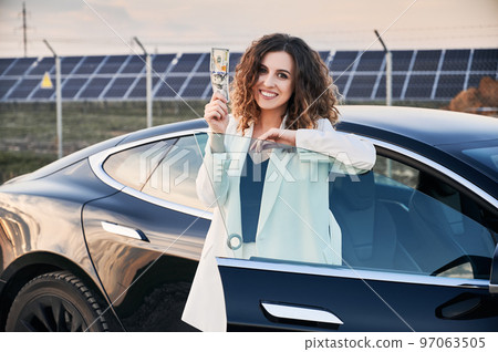 Portrait of business lady near open car, holding banknote against solar power plant. Female owner of ecological auto with electric motor demonstrating budget savings when using electric vehicles. Portrait of business lady near open car, holding banknote against solar power plant. Female owner of ecological auto with electric motor demonstrating budget savings when using electric vehicles. 97063505
