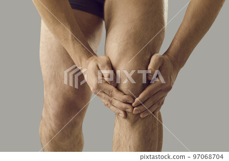 Close up of hands of man holding and massaging sore knee standing on gray background. 97068704