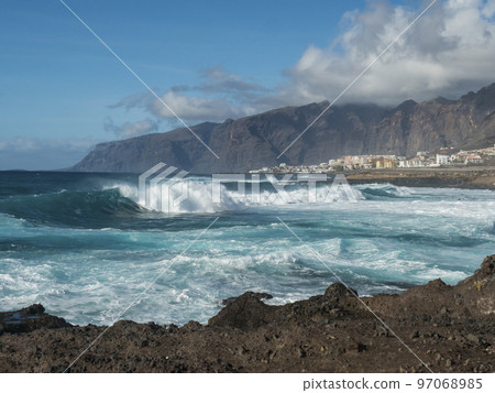 Big breaking waves splash against black lava rock coast. Turquoise blue Atlantic ocean with view of Los Gigantes cliffs and village. Tenerife, Canary islands, Spain. Sunny winter day. Big breaking waves splash against black lava rock coast. Turquoise blue Atlantic ocean with view of Los Gigantes cliffs and village. Tenerife, Canary islands, Spain. Sunny winter day. 97068985