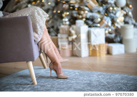 Women's legs. Cropped close-up photo of healthy beautiful elegant female legs in beige high-heeled shoes, woman sitting in a chair on the background of a Christmas tree with gifts Women's legs. Cropped close-up photo of healthy beautiful elegant female legs in beige high-heeled shoes, woman sitting in a chair on the background of a Christmas tree with gifts 97070380