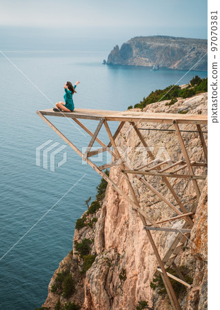 A girl sits on a wooden springboard for jumping with a rope. In a dark green dress with her hands in the air. A girl sits on a wooden springboard for jumping with a rope. In a dark green dress with her hands in the air. 97070381