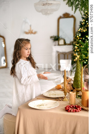 The girl helps to set the table on Christmas Eve, placing plates on a decorated table with a tablecloth. Preparation for the festive celebration of the new year The girl helps to set the table on Christmas Eve, placing plates on a decorated table with a tablecloth. Preparation for the festive celebration of the new year 97073406