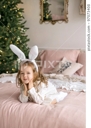 A beautiful girl with a headdress with rabbit ears in a white dress lies on the bed with her cheeks pressed with her hands. Christmas decorated Christmas tree in the bedroom A beautiful girl with a headdress with rabbit ears in a white dress lies on the bed with her cheeks pressed with her hands. Christmas decorated Christmas tree in the bedroom 97073408