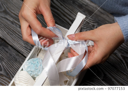 Marshmallow cones. Homemade zephyr. Packed in a wooden decorative box. A woman ties a ribbon into a bow. On the background of the boards. Close-up. 97077153