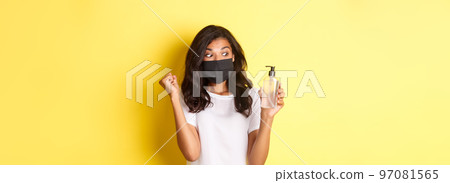 Concept of covid-19, social distancing and lifestyle. Image of happy african-american woman in face mask and white t-shirt, showing good hand sanitizer and making a fist pump, yellow background 97081565