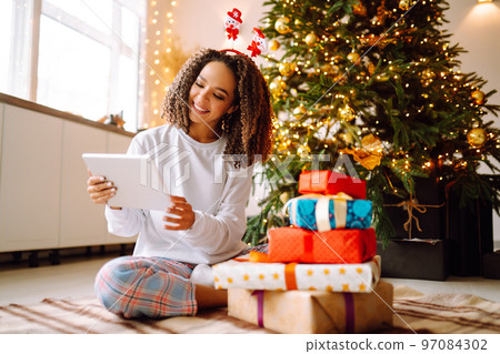 A young woman on the background of a Christmas tree with gifts, with a tablet has a video call or video chat. 97084302