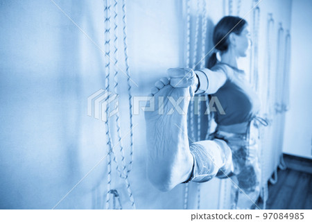 Young girl doing yoga pose holding her toe standing against wall with ropes 97084985