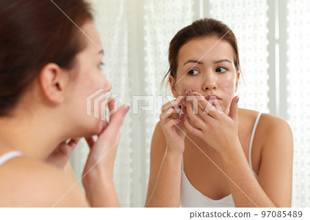Teen girl with acne problem squeezing pimple near mirror in bathroom Teen girl with acne problem squeezing pimple near mirror in bathroom 97085489