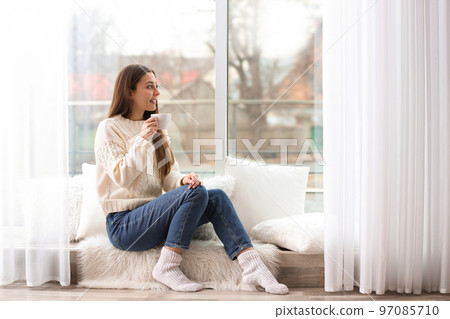 Young woman with cup near window indoors Young woman with cup near window indoors 97085710