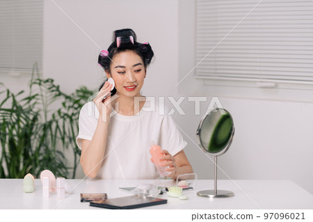 Beauty Routine. Attractive young female applying makeup removal milk on cotton pad while standing near mirror 97096021