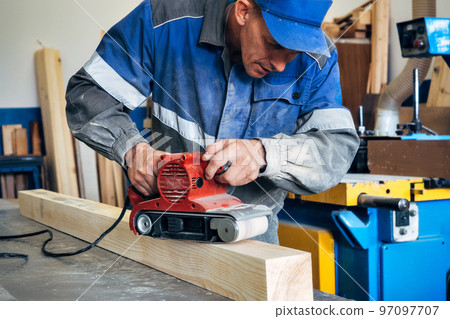 Carpenter in work clothes processes wood with grinder. Polishing of wood surface. Middle-aged man 97097707