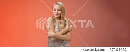 Tender feminine elegant blond young woman in stylish silver glittering dress close eyes dreamy smiling touching herself cuddle embrace hands crossed body remember heartwarming passionate feelings Tender feminine elegant blond young woman in stylish silver glittering dress close eyes dreamy smiling touching herself cuddle embrace hands crossed body remember heartwarming passionate feelings 97101082