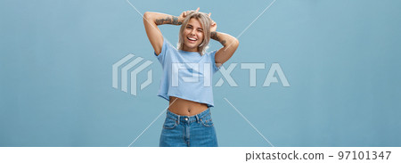 Studio shot of cute attractive and joyful stylish blond woman with tattoos on arms winking and smiling broadly holding index fingers on head making horns standing in summer outfit over blue wall Studio shot of cute attractive and joyful stylish blond woman with tattoos on arms winking and smiling broadly holding index fingers on head making horns standing in summer outfit over blue wall 97101347