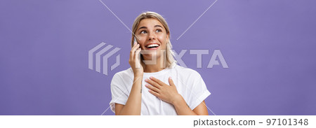 Waist-up shot of thankful charming blonde female student with tanned skin and blond hair holding hand on breast receiving delightful warm call from loving friend holding smartphone near ear gazing up Waist-up shot of thankful charming blonde female student with tanned skin and blond hair holding hand on breast receiving delightful warm call from loving friend holding smartphone near ear gazing up 97101348